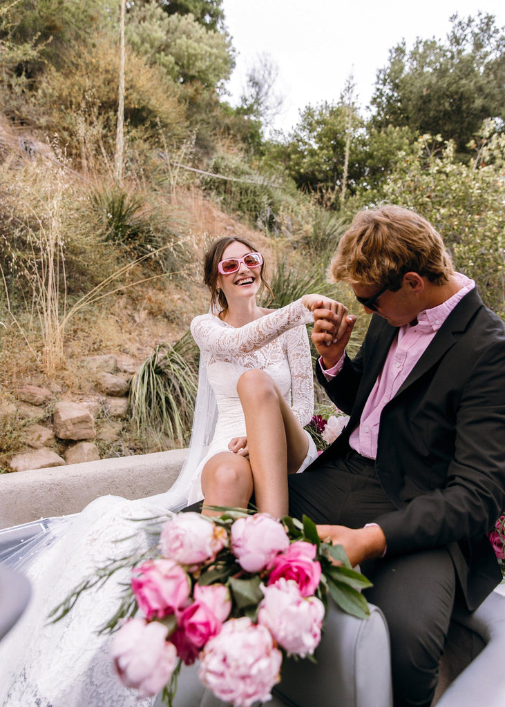 Bride in pink sunglasses and Sasha dress playing with groom in back of white convertible