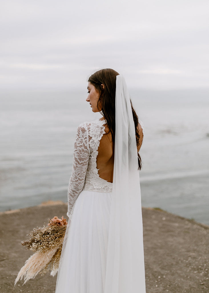 Bride standing on a cliff overlooking the ocean wearing Kenna chiffon veil and Indy dress, from behind