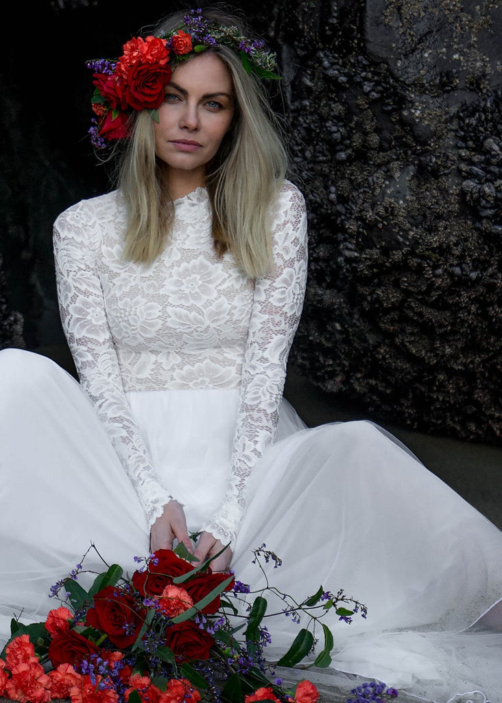 Bride with vibrant flower crown and bouquet wearing Zoey 2.0 wedding dress sitting by rocks