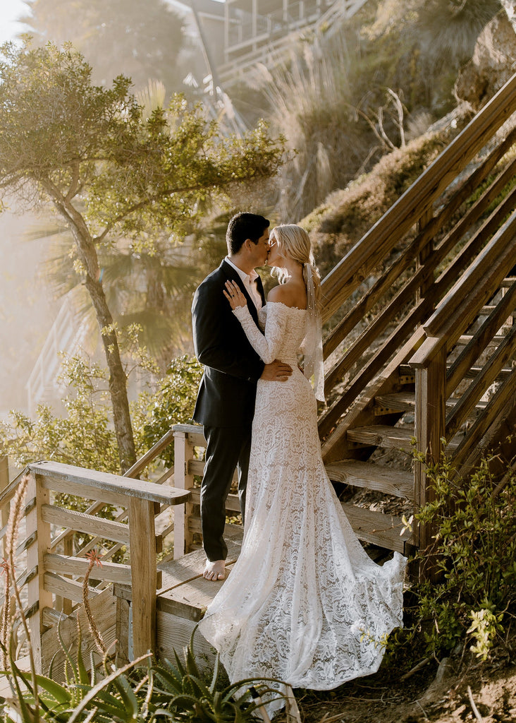 Groom kissing blonde bride wearing Ayla dress on outdoor stairs leading to beach 