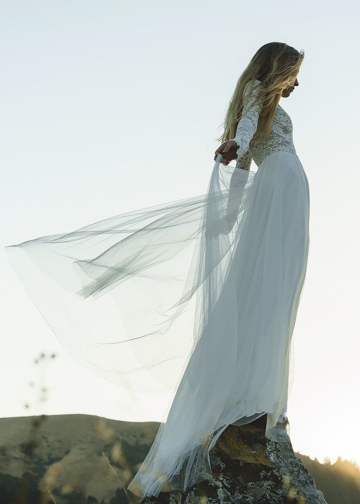 Bride wearing Zoey 2.0 dress standing on top of a rock, holding her arms out, looking away from the camera