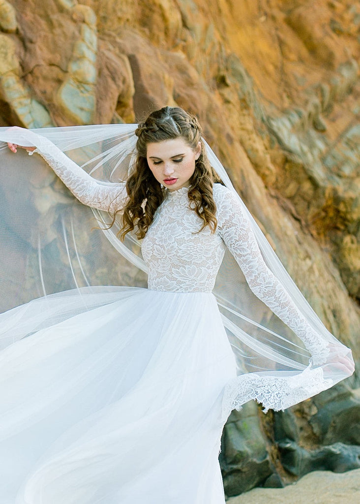 Bride in veil wearing Zoey 2.0 dress standing arms out on beach by rocks