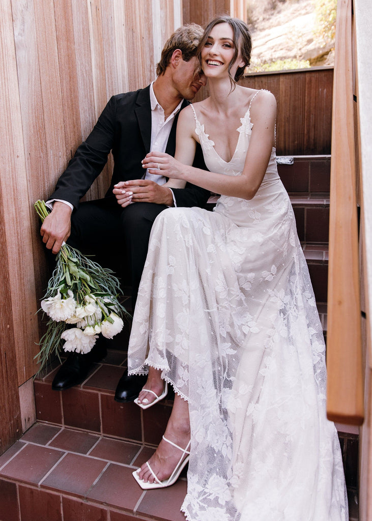 Bride wearing Lilith Dress sitting on stairs with groom, smiling