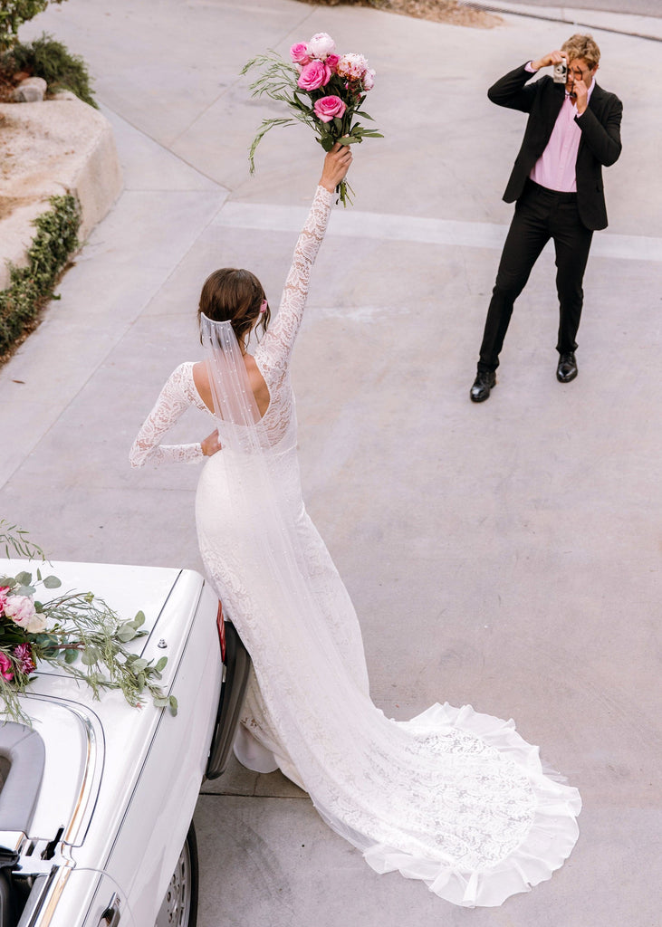 Bride wearing Sasha Dress holding up bouquet in front of white convertible as groom photographs her