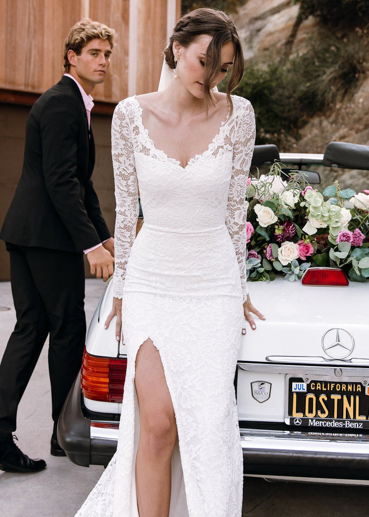 Bride wearing Sia Dress leaning against white convertible, groom looking at her in the background