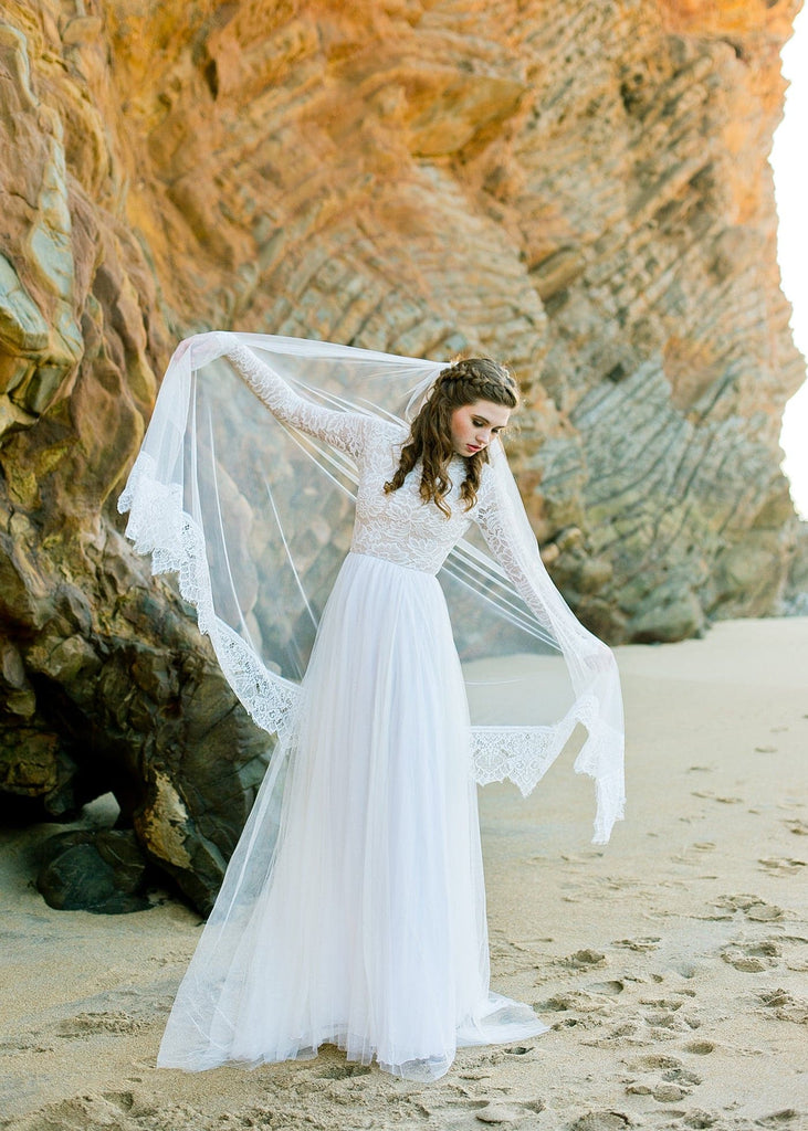 Bride wearing veil and Zoey 2.0 dress standing on beach by large rock formation